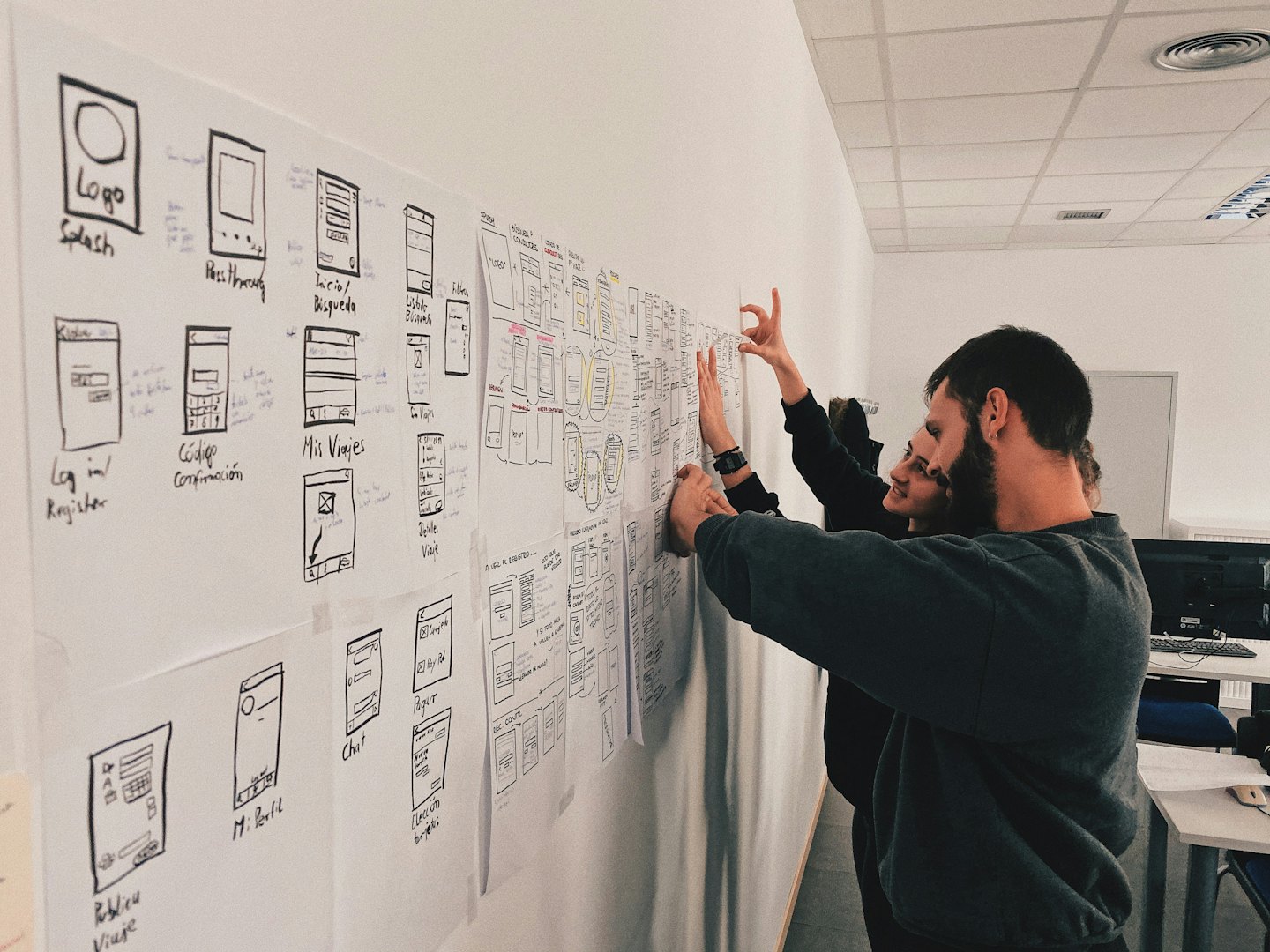 Two people collaborating on UI/UX wireframes taped to an office wall.